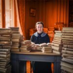 Vrouw zit aan een bureau omringd door hoge stapels boeken, verwijzend naar kennis, normering en intellectuele arbeid.