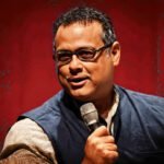 Vijay Prashad spreekt in een zaal terwijl hij een microfoon vasthoudt, met geconcentreerde blik richting publiek.