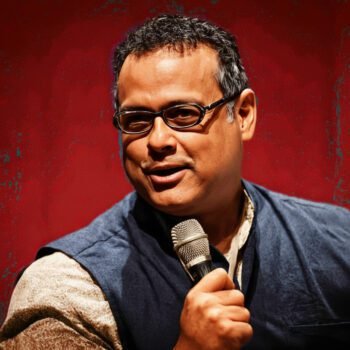 Vijay Prashad spreekt in een zaal terwijl hij een microfoon vasthoudt, met geconcentreerde blik richting publiek.