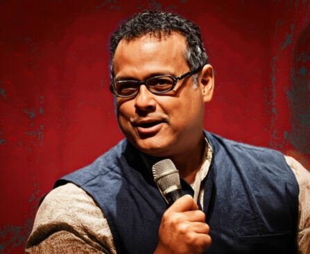 Vijay Prashad spreekt in een zaal terwijl hij een microfoon vasthoudt, met geconcentreerde blik richting publiek.