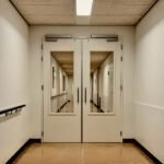 Dubbele deuren in een zorginstelling die toegang geven tot een lange, lege gang, symbool voor institutionele macht en normalisering.