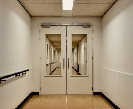 Dubbele deuren in een zorginstelling die toegang geven tot een lange, lege gang, symbool voor institutionele macht en normalisering.