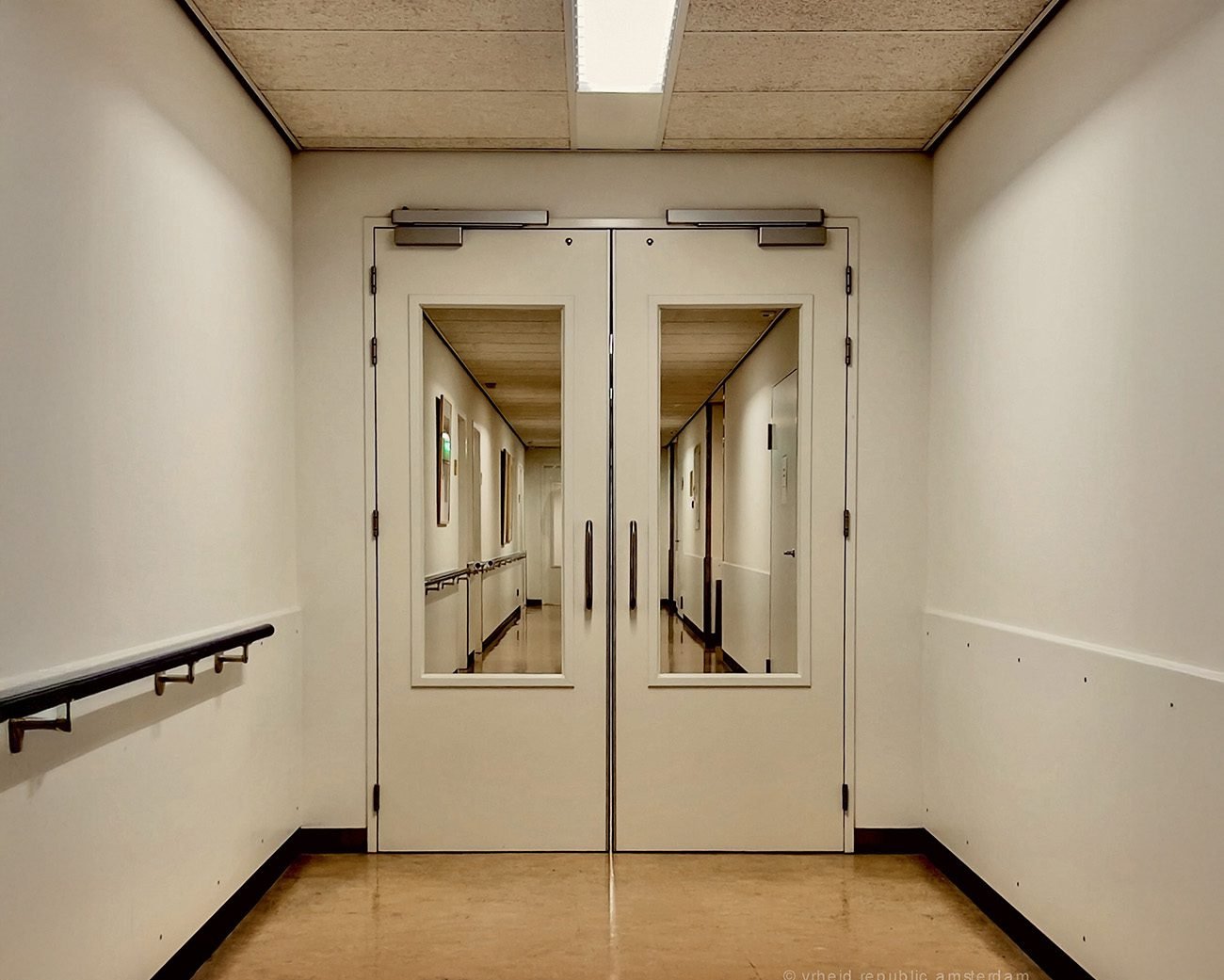 Dubbele deuren in een zorginstelling die toegang geven tot een lange, lege gang, symbool voor institutionele macht en normalisering.