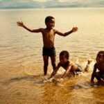 Zwarte kinderen spelen in ondiep water aan een kust of rivier in de Verenigde Staten, een plek waar families generaties lang samenkwamen en kinderen leerden zwemmen.