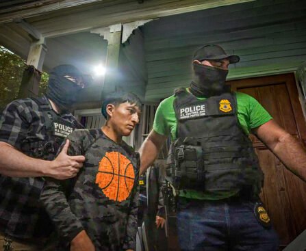 Gemaskerde agenten van U.S. Customs and Border Protection arresteren een man bij een woning terwijl zij hem vasthouden aan beide armen.