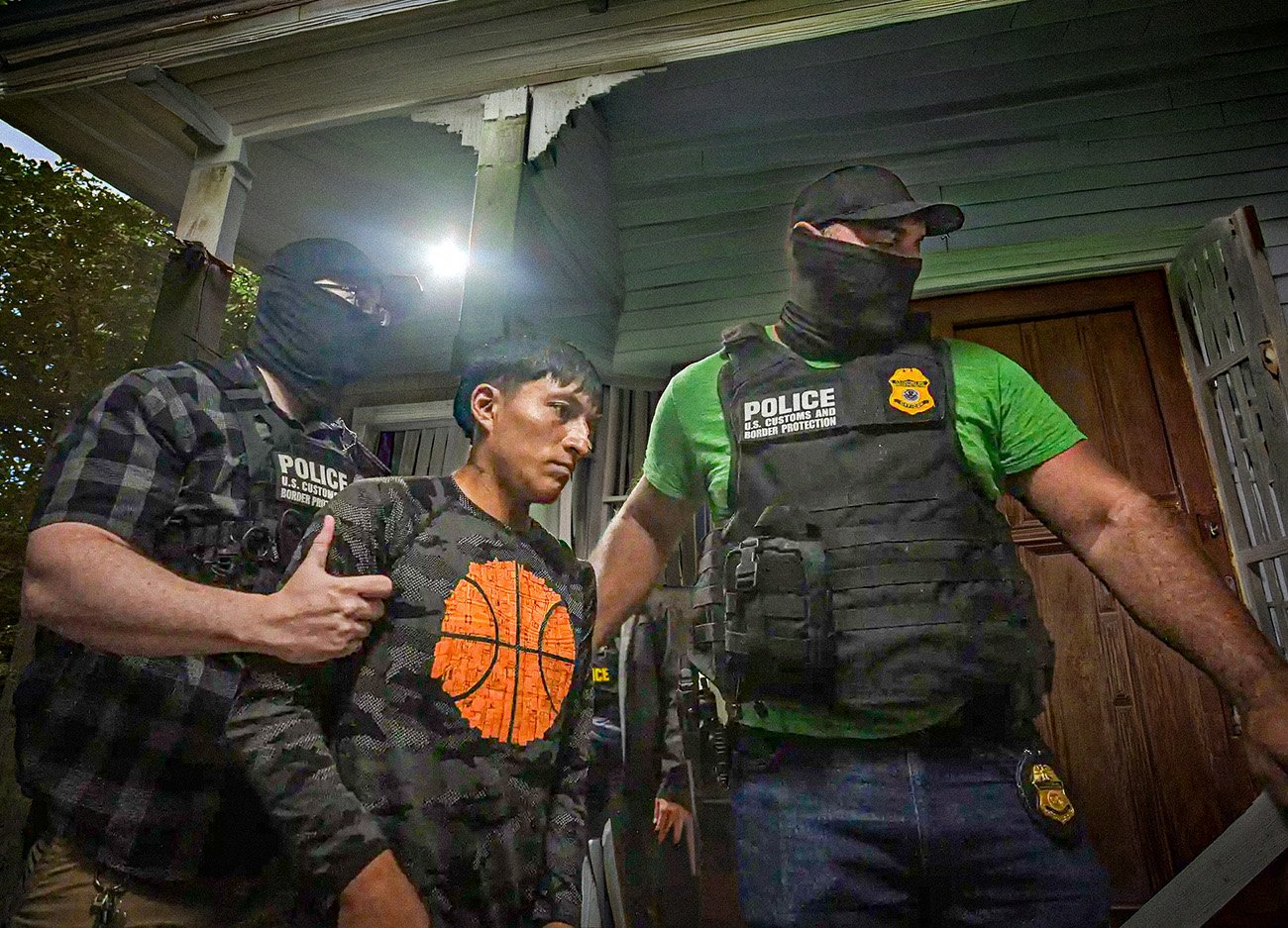 Gemaskerde agenten van U.S. Customs and Border Protection arresteren een man bij een woning terwijl zij hem vasthouden aan beide armen.