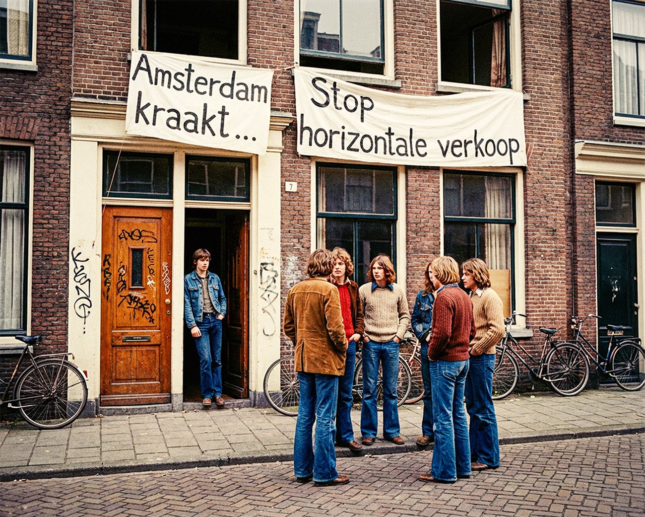 Jonge activisten staan voor een Amsterdams woonhuis met spandoeken “Amsterdam kraakt” en “Stop horizontale verkoop”, symbool van de kraakbeweging en de strijd om betaalbare huisvesting.