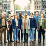 Groep verschillende Amsterdammers staat samen op een brug in Amsterdam, met grachtenpanden, fietsen en een tram op de achtergrond.