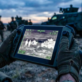 Handen in militaire handschoenen houden een dronecontroller met thermisch beeld en doelmarkeringen tijdens een veldoperatie.