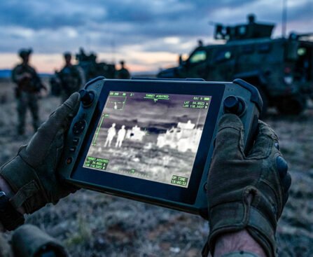Handen in militaire handschoenen houden een dronecontroller met thermisch beeld en doelmarkeringen tijdens een veldoperatie.