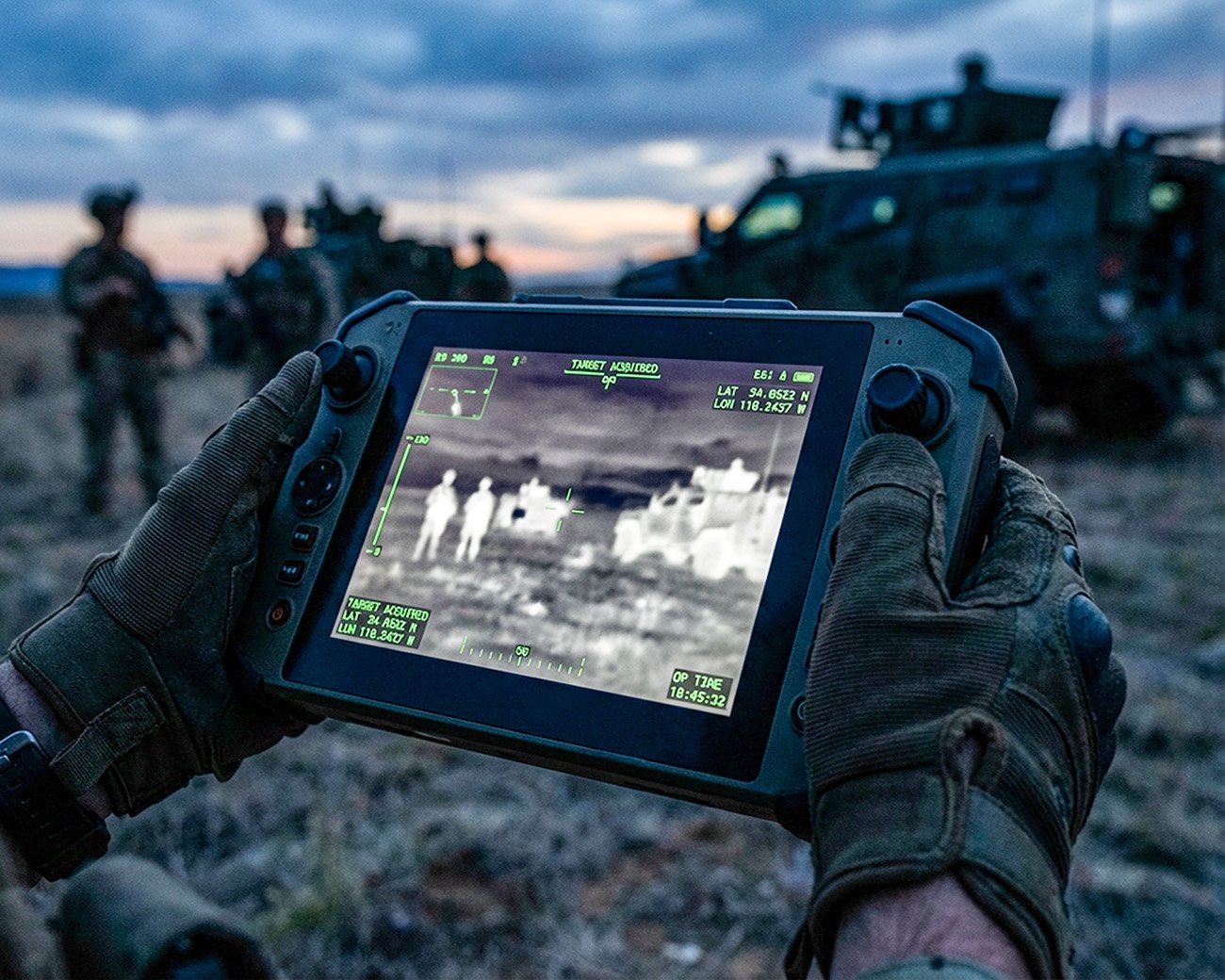 Handen in militaire handschoenen houden een dronecontroller met thermisch beeld en doelmarkeringen tijdens een veldoperatie.
