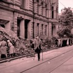 Mensen ruimen puin in een verwoeste Duitse stad na de Tweede Wereldoorlog terwijl een man met krukken door de straat loopt