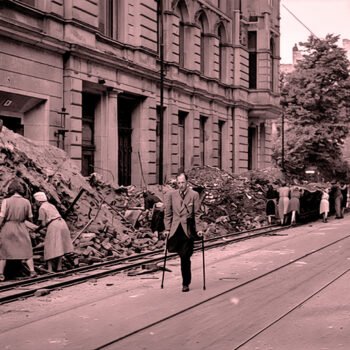 Mensen ruimen puin in een verwoeste Duitse stad na de Tweede Wereldoorlog terwijl een man met krukken door de straat loopt
