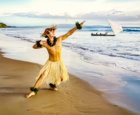 Persoon in traditionele kledij aan de kust van Guam met een zeilkano op zee, als verwijzing naar CHamoru cultuur, zeevaart en zelfbeschikking.