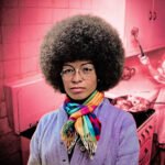 Portret van een vrouw op de voorgrond, met op de achtergrond een keuken en huiselijk werk als verwijzing naar zorg, huiswerk en kapitalisme.