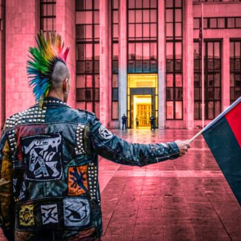 Een demonstrant met een rood-zwarte vlag staat voor een verlicht overheidsgebouw, terwijl de spanning tussen protest, autonomie en institutionele macht zichtbaar wordt.