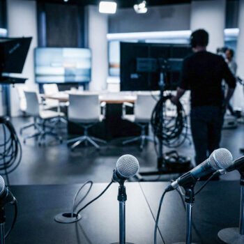Microfoons op een tafel in een televisiestudio, met op de achtergrond een debatset en studiopersoneel.