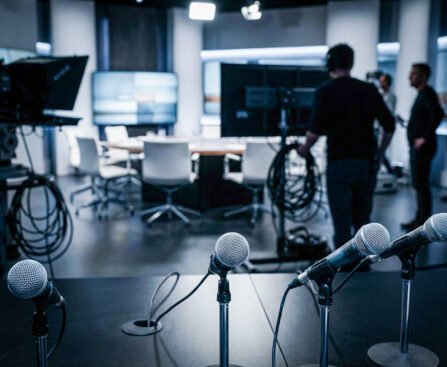 Microfoons op een tafel in een televisiestudio, met op de achtergrond een debatset en studiopersoneel.