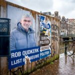 Gescheurde poster van Rob Oudkerk op een houten wand langs een Amsterdamse gracht met fietsen, graffiti en natte straatstenen.