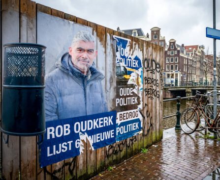 Gescheurde poster van Rob Oudkerk op een houten wand langs een Amsterdamse gracht met fietsen, graffiti en natte straatstenen.