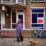 Vrouw met koffer staat voor een Amsterdamse woning met een te koop-bord in het raam, naast een fiets en een hond op straat.