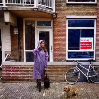 Vrouw met koffer staat voor een Amsterdamse woning met een te koop-bord in het raam, naast een fiets en een hond op straat.