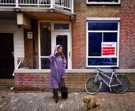 Vrouw met koffer staat voor een Amsterdamse woning met een te koop-bord in het raam, naast een fiets en een hond op straat.