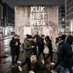 Mensen lopen langs een natte stadsstraat terwijl op een betonnen muur “Kijk niet weg” staat.