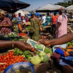 Handen wisselen CFA-frankbiljetten uit op een Afrikaanse markt, tussen groente, stoffen en marktkramen.