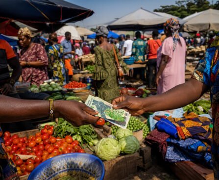 Handen wisselen CFA-frankbiljetten uit op een Afrikaanse markt, tussen groente, stoffen en marktkramen.