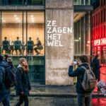 Mensen lopen langs een glazen vergaderruimte terwijl op een betonnen muur “Ze zagen het wel” staat.