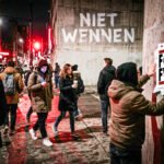 Mensen lopen door een donkere stadsstraat terwijl op een muur “Niet wennen” staat en iemand een poster tegen FVD ophangt.
