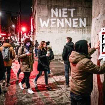 Mensen lopen door een donkere stadsstraat terwijl op een muur “Niet wennen” staat en iemand een poster tegen FVD ophangt.