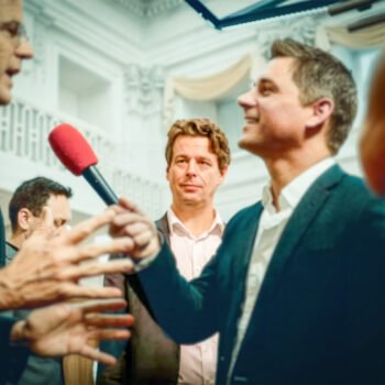 Journalist houdt een microfoon bij politici en omstanders in een politieke omgeving, tijdens een gesprek over media en macht.