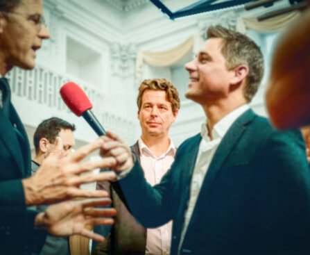 Journalist houdt een microfoon bij politici en omstanders in een politieke omgeving, tijdens een gesprek over media en macht.
