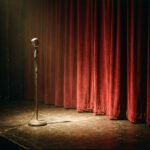 Microfoon op een leeg podium voor een gesloten rood gordijn, als beeld voor literatuur, weigering en de normalisering van geweld.