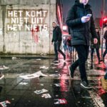 Mensen lopen over een natte stadsstraat terwijl op een muur “Het komt niet uit het niets” staat.
