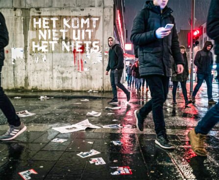 Mensen lopen over een natte stadsstraat terwijl op een muur “Het komt niet uit het niets” staat.