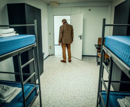 Een man staat met zijn rug naar de camera in een sobere slaapruimte met stapelbedden in een opvanglocatie.