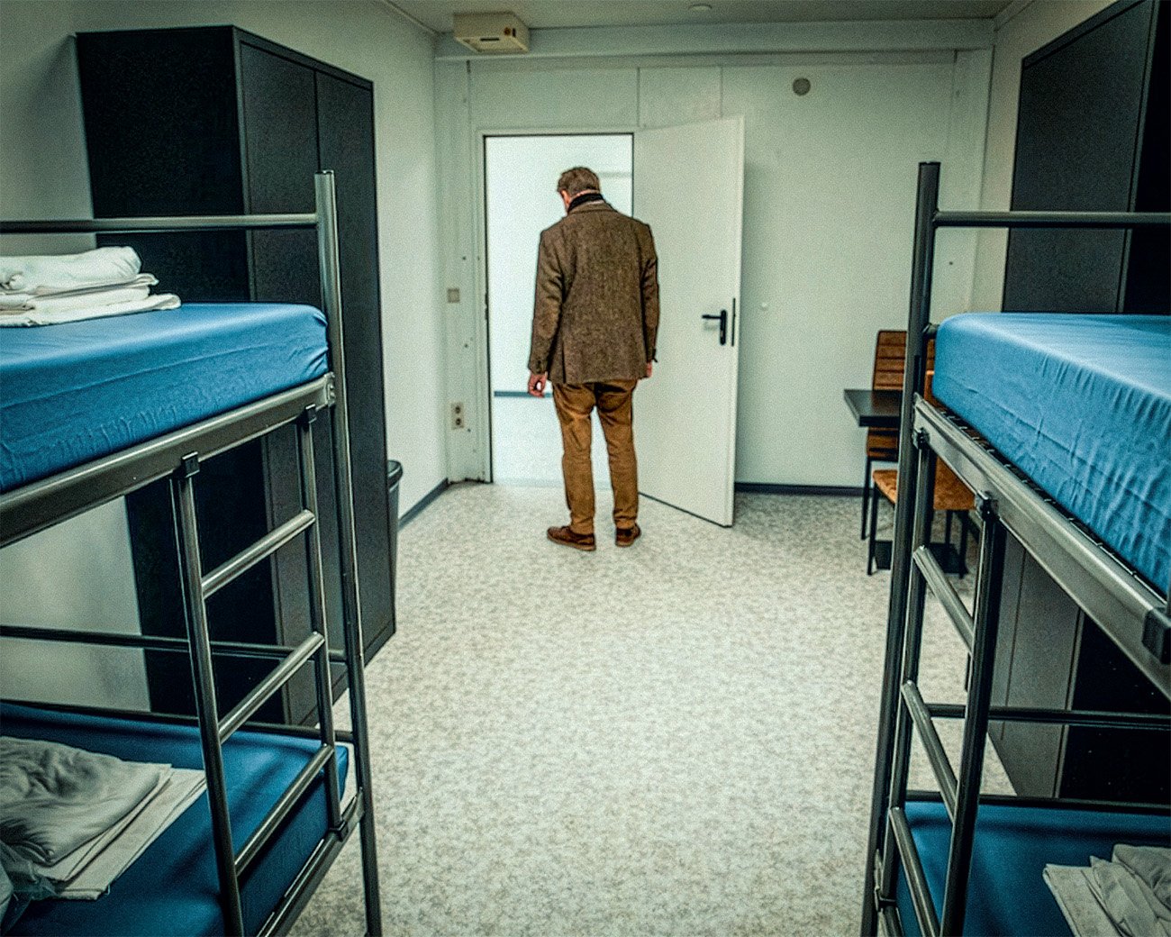 Een man staat met zijn rug naar de camera in een sobere slaapruimte met stapelbedden in een opvanglocatie.