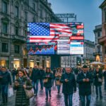 Europees stadsplein met mensen op telefoons onder een groot digitaal scherm met Amerikaanse vlag, nieuwsflitsen en beursgrafieken.
