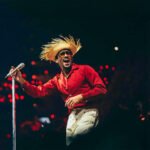 Bad Bunny staat op een groot podium voor een tropisch decor met palmen, zee en felblauwe lucht.