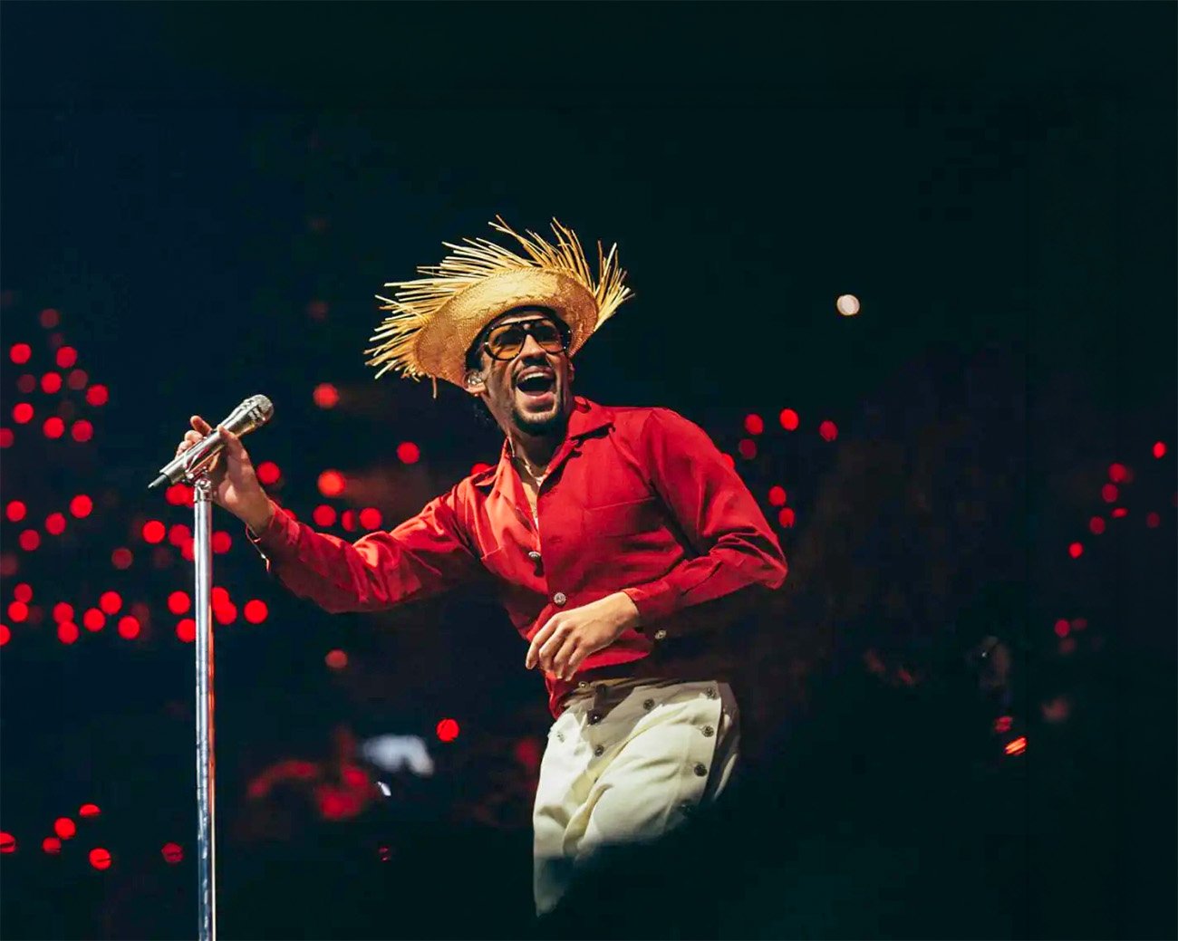 Bad Bunny staat op een groot podium voor een tropisch decor met palmen, zee en felblauwe lucht.