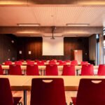 Lege zaal met rode stoelen, tafels en een projectiescherm waar geen spreker aanwezig is.