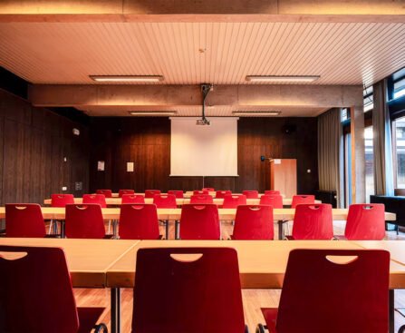 Lege zaal met rode stoelen, tafels en een projectiescherm waar geen spreker aanwezig is.