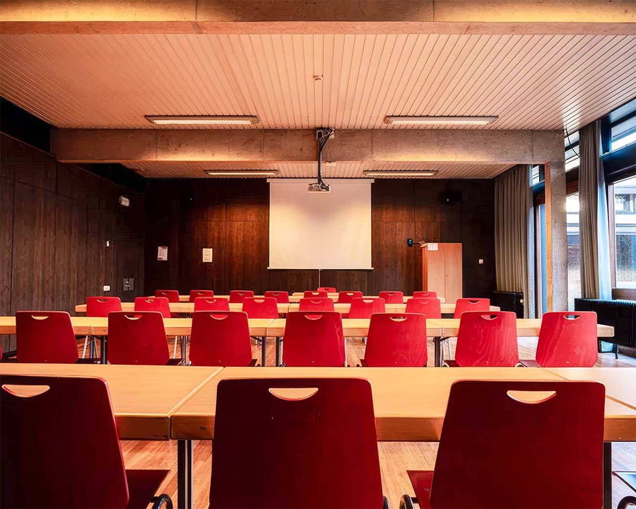 Lege zaal met rode stoelen, tafels en een projectiescherm waar geen spreker aanwezig is.