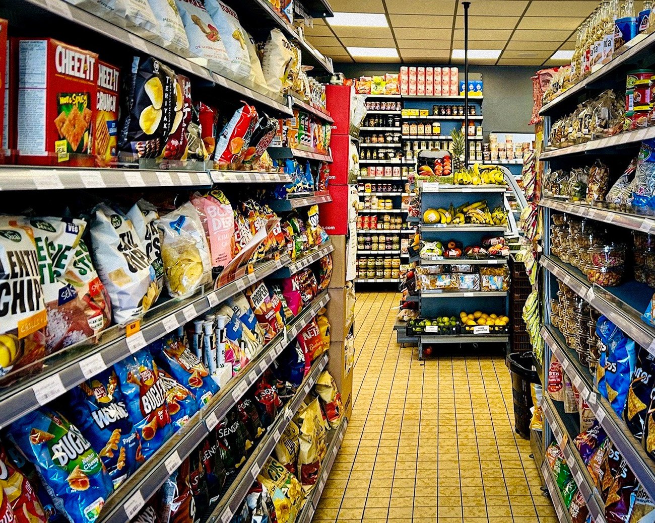 Supermarktpad vol zakken chips, snacks en bewerkte voeding, met een kleine fruit- en groenteafdeling op de achtergrond.