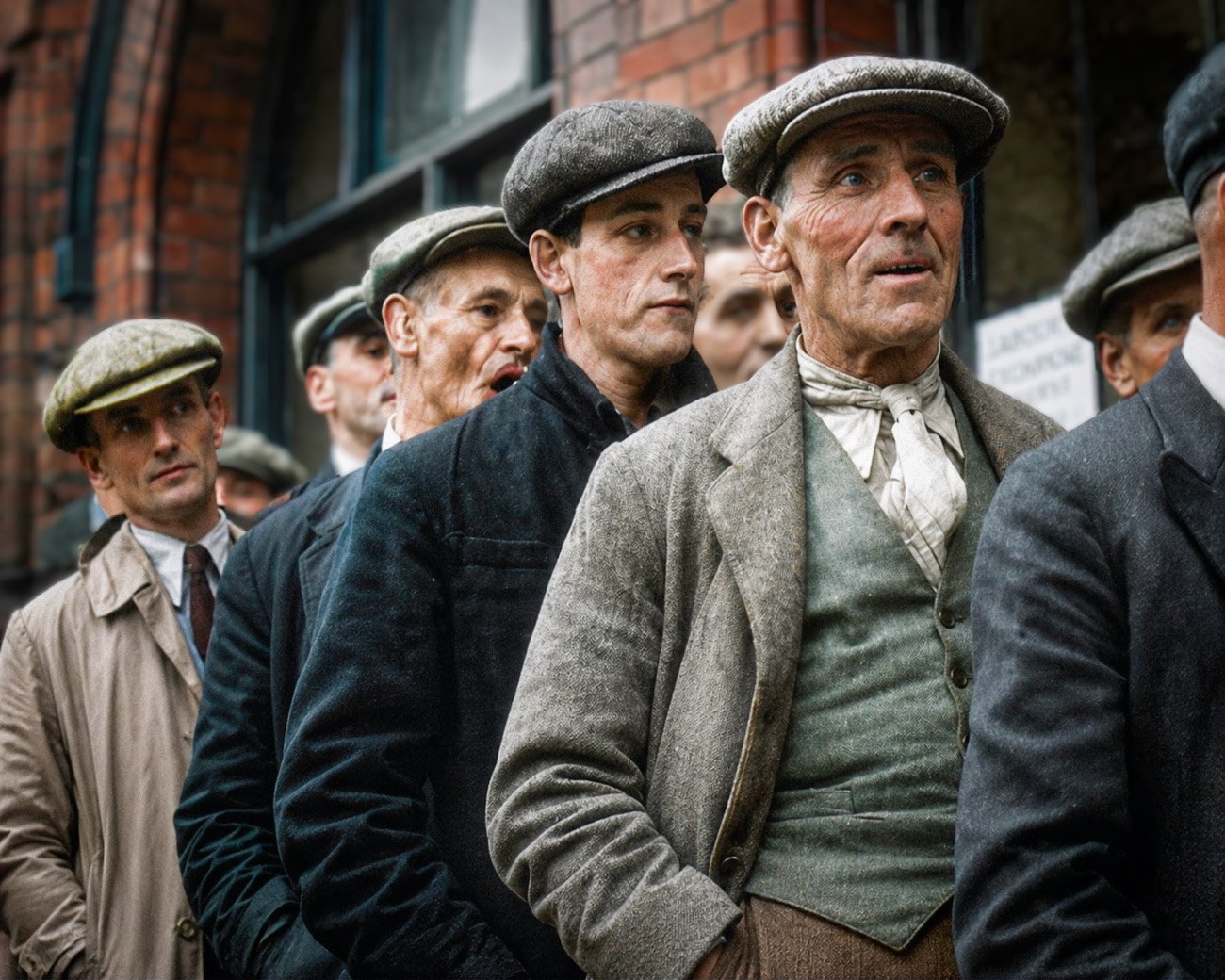 Groep arbeiders met petten en jassen staat dicht op elkaar bij een gebouw in het East End rond 1930.