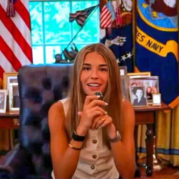 Jonge vrouw zit achter het presidentiële bureau in een Oval Office-achtige ruimte met Amerikaanse vlaggen, familiefoto’s en staatsdecor op de achtergrond.
