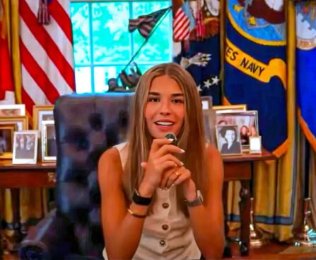 Jonge vrouw zit achter het presidentiële bureau in een Oval Office-achtige ruimte met Amerikaanse vlaggen, familiefoto’s en staatsdecor op de achtergrond.